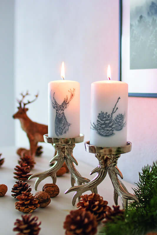 Zwei Weihnachtskerzen mit Hirsch- und Tannenzapfenmotiv in gemütlicher Tischdekoration mit Naturmaterialien.